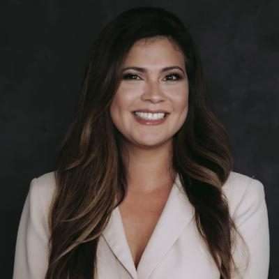Professional headshot of a smiling woman in a light-colored blazer against a dark studio background.